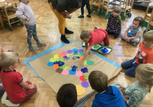 Dzieci przyklejają kolorowe kółka na szary papier tworząc stonogę.